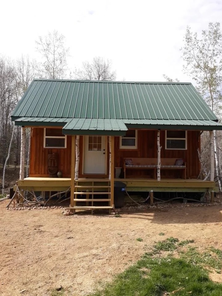 A finished cabin with a green metal roof standing on bare graded dirt, nothing planted around it